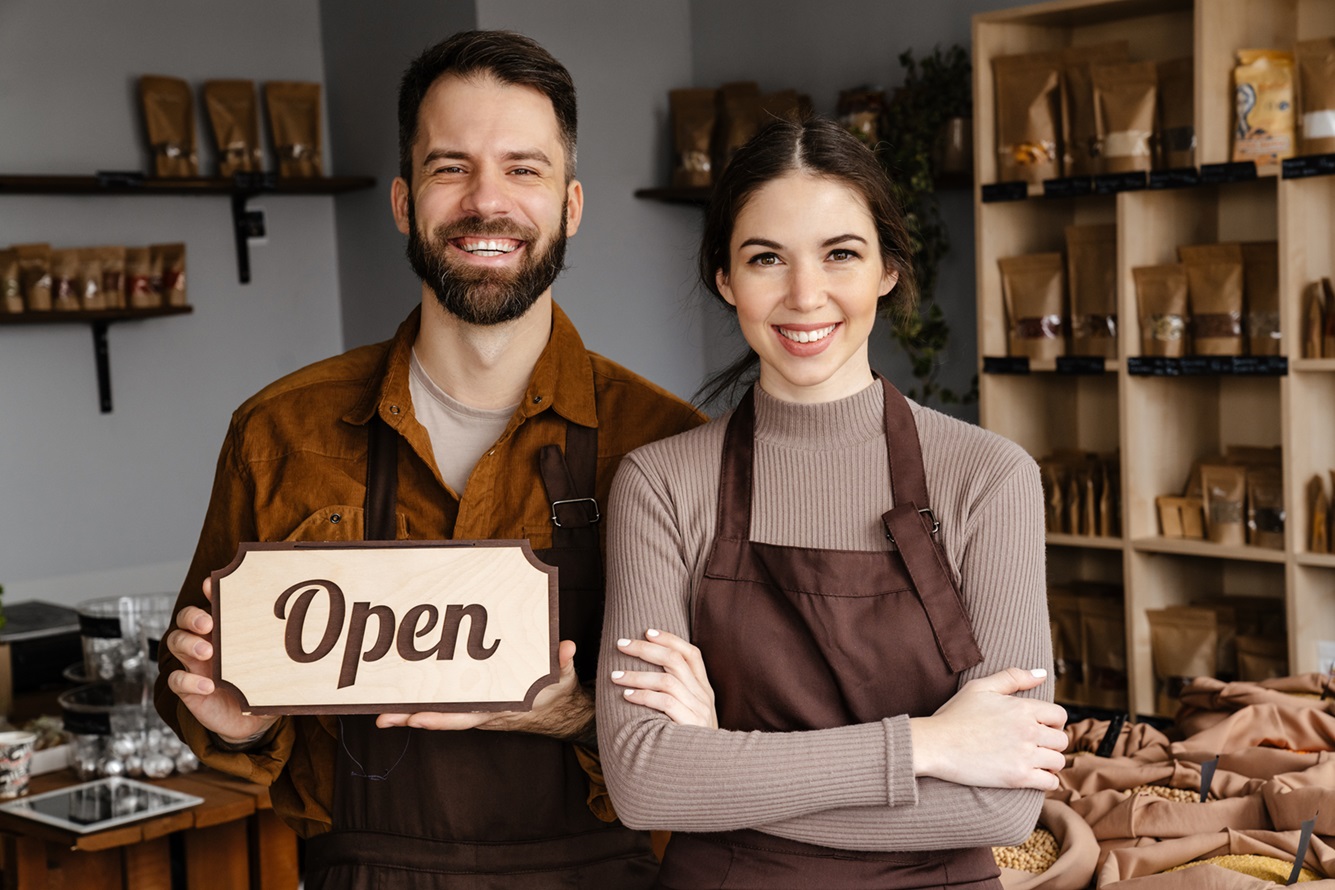 Smiling sellers man and woman in aprons showing open sign board at local eco shop